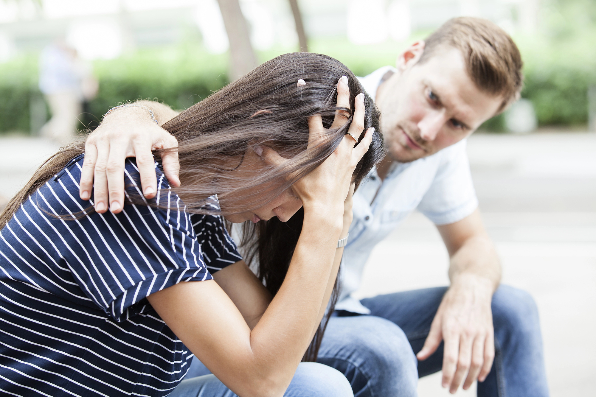 A man comforts a distressed woman sitting beside him. The woman, dressed in a striped shirt, has her head in her hands. The man, in a light-colored shirt and jeans, gently places his hand on her shoulder, expressing concern and support.