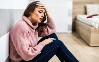 A woman with long brown hair and wearing a pink sweater and blue jeans sits on the floor against a wall, looking distressed. She rests her forehead on her hand and has an expression of concern, with a bed and wardrobe visible in the background.