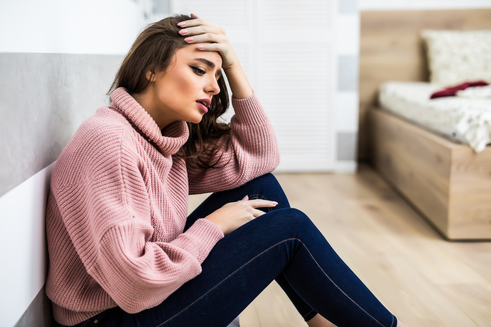 A woman with long brown hair and wearing a pink sweater and blue jeans sits on the floor against a wall, looking distressed. She rests her forehead on her hand and has an expression of concern, with a bed and wardrobe visible in the background.