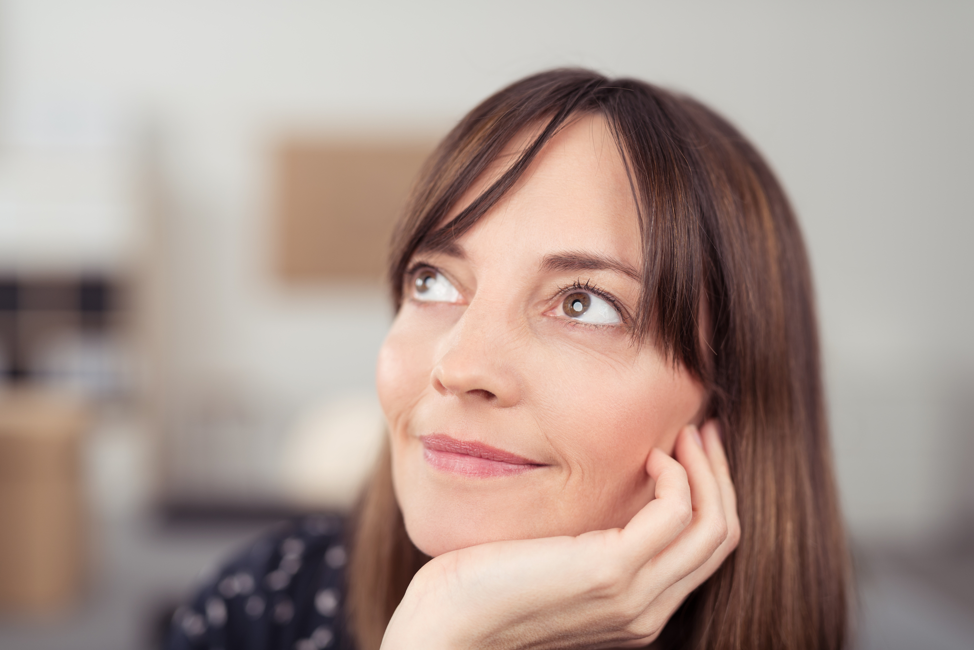 A woman with light skin and straight brown hair rests her chin on her hand while looking upward with a thoughtful expression. She is indoors, and the background is softly blurred.