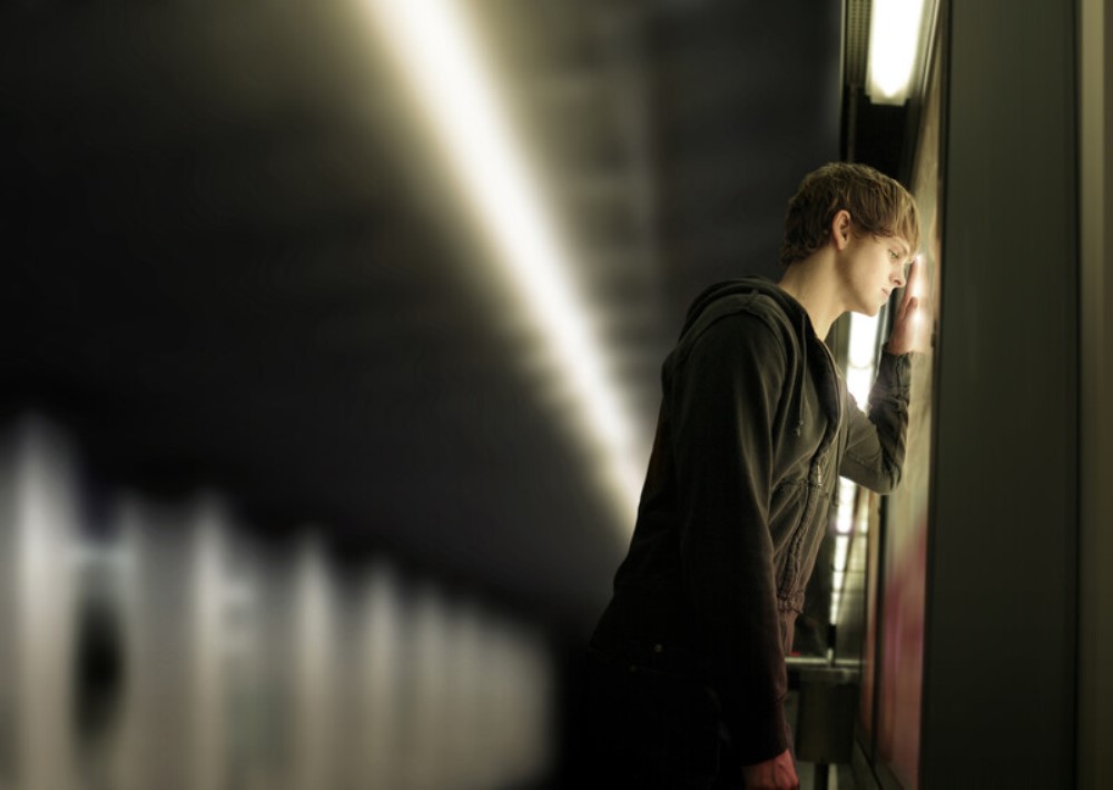 A person with short hair wearing a dark hoodie leans against a glass wall with their forearm while looking down, creating a pensive or contemplative pose. The background is blurred, giving the impression of a corridor or tunnel.