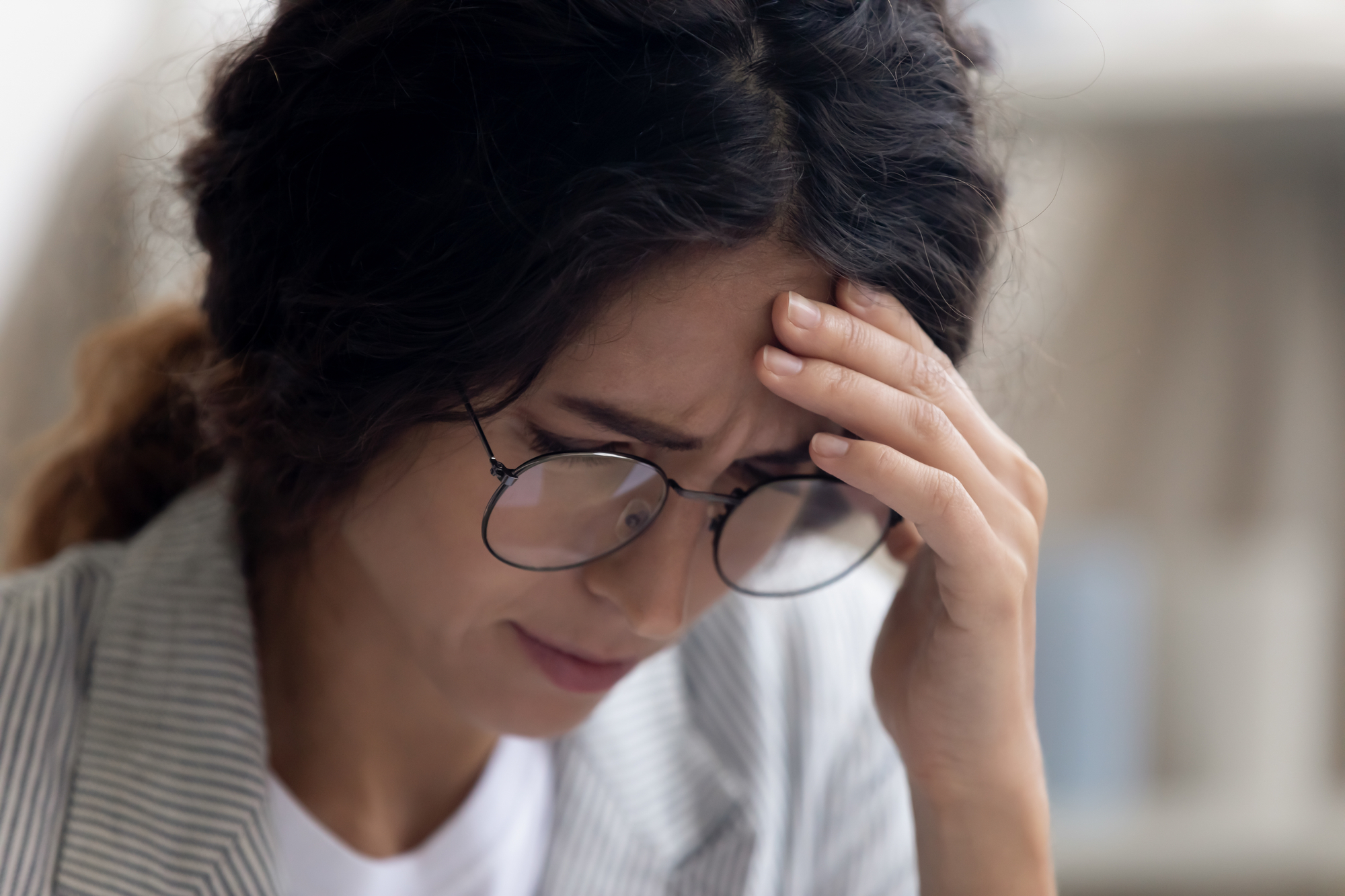 A woman with long dark hair and wearing glasses holds her forehead with a hand, appearing stressed or worried. She has a serious expression and is dressed in a light-colored blazer. The background is blurred.