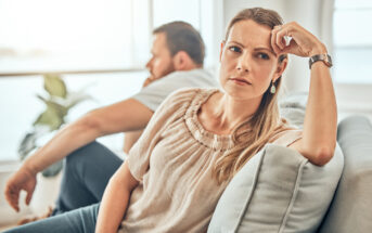 A woman with long blonde hair wearing a beige top sits on a sofa with a pensive expression, resting her head on her hand. Behind her, a man with short hair and a beard, wearing a white t-shirt, sits turned away, seemingly deep in thought.