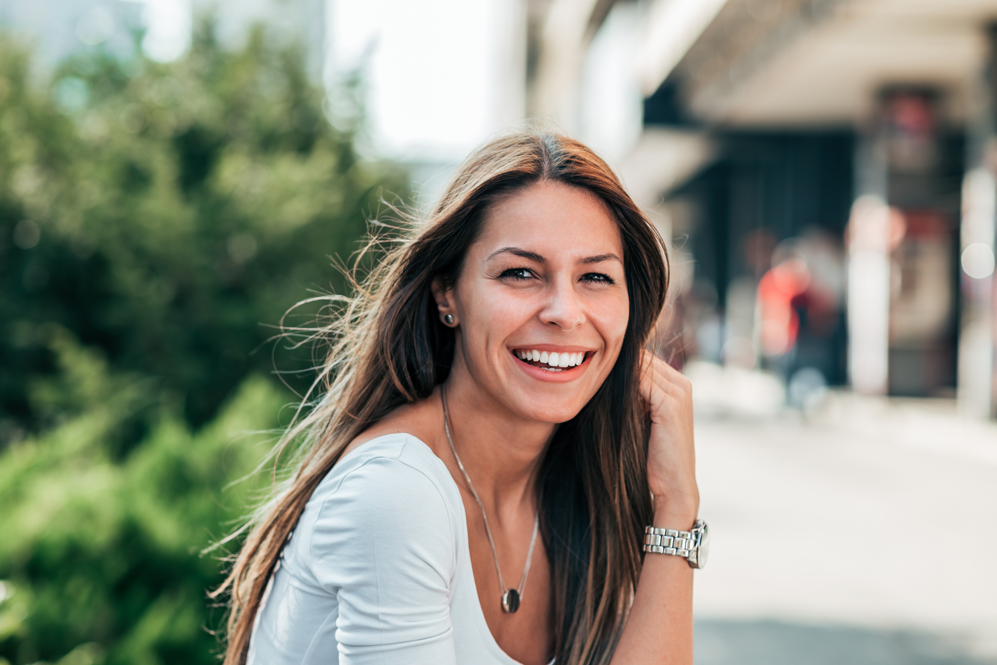 A woman with long brown hair is smiling at the camera while sitting outside on a sunny day. She is wearing a white top, a silver watch, and a necklace. The background is slightly blurred, showing green foliage and some buildings.