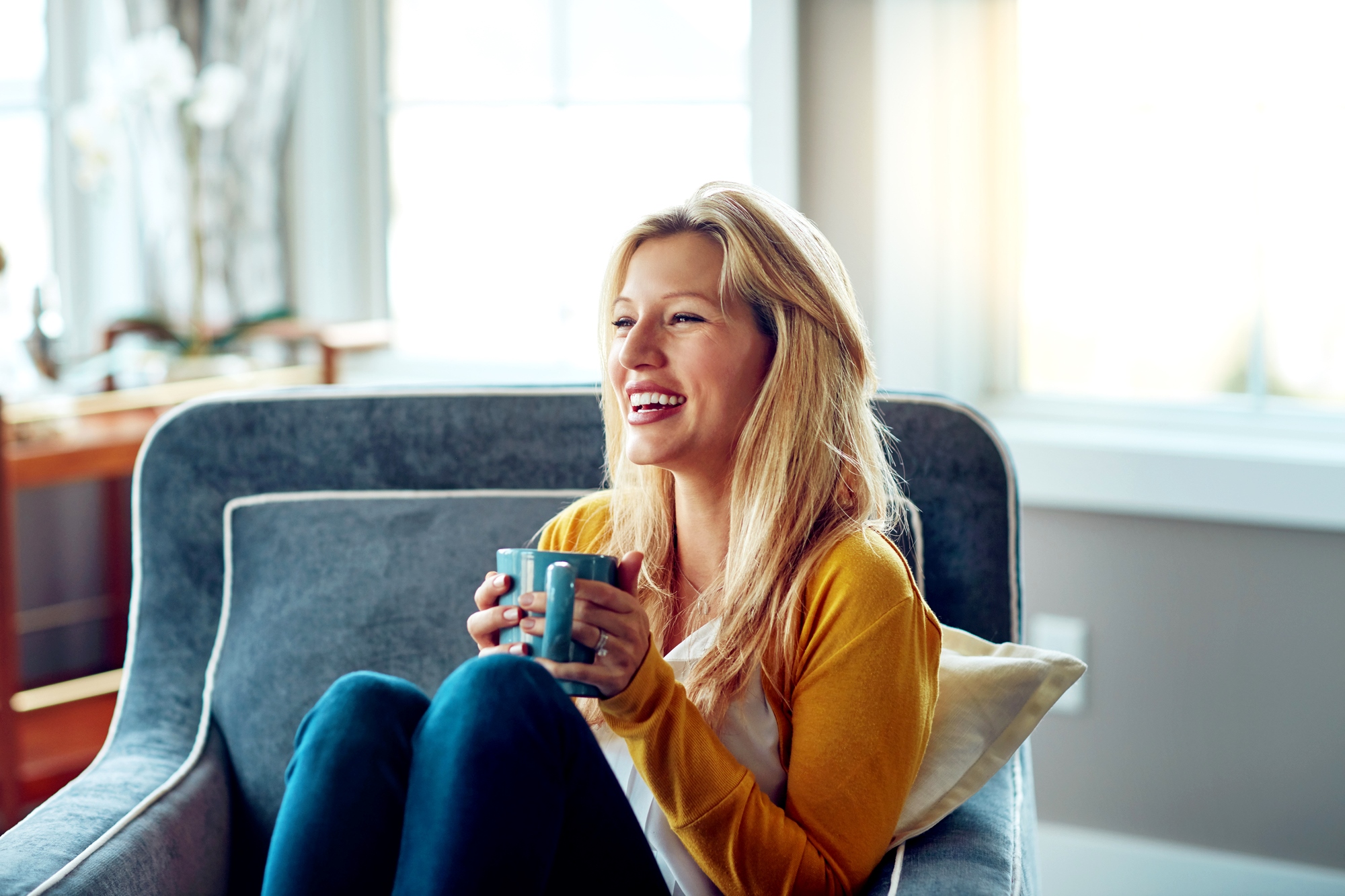 A woman with long blonde hair, wearing a yellow sweater and blue jeans, sits comfortably on a gray couch, holding a blue mug and smiling. Sunlight shines through the windows in the background, illuminating the room.