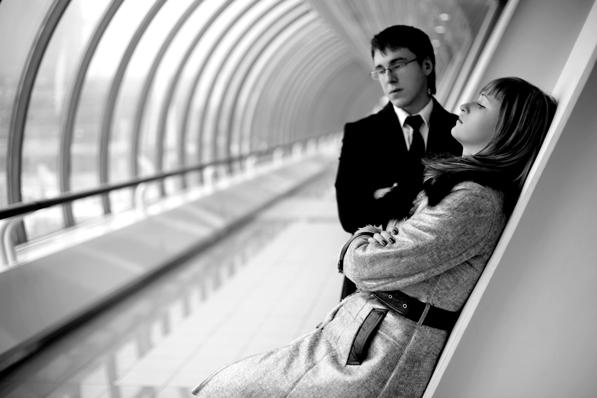 A black and white image of two people inside a modern, arched hallway. A woman wearing a coat leans against a wall with her arms crossed and eyes closed. A man in a suit, also with arms crossed, stands nearby looking at her. The corridor features large window panes.