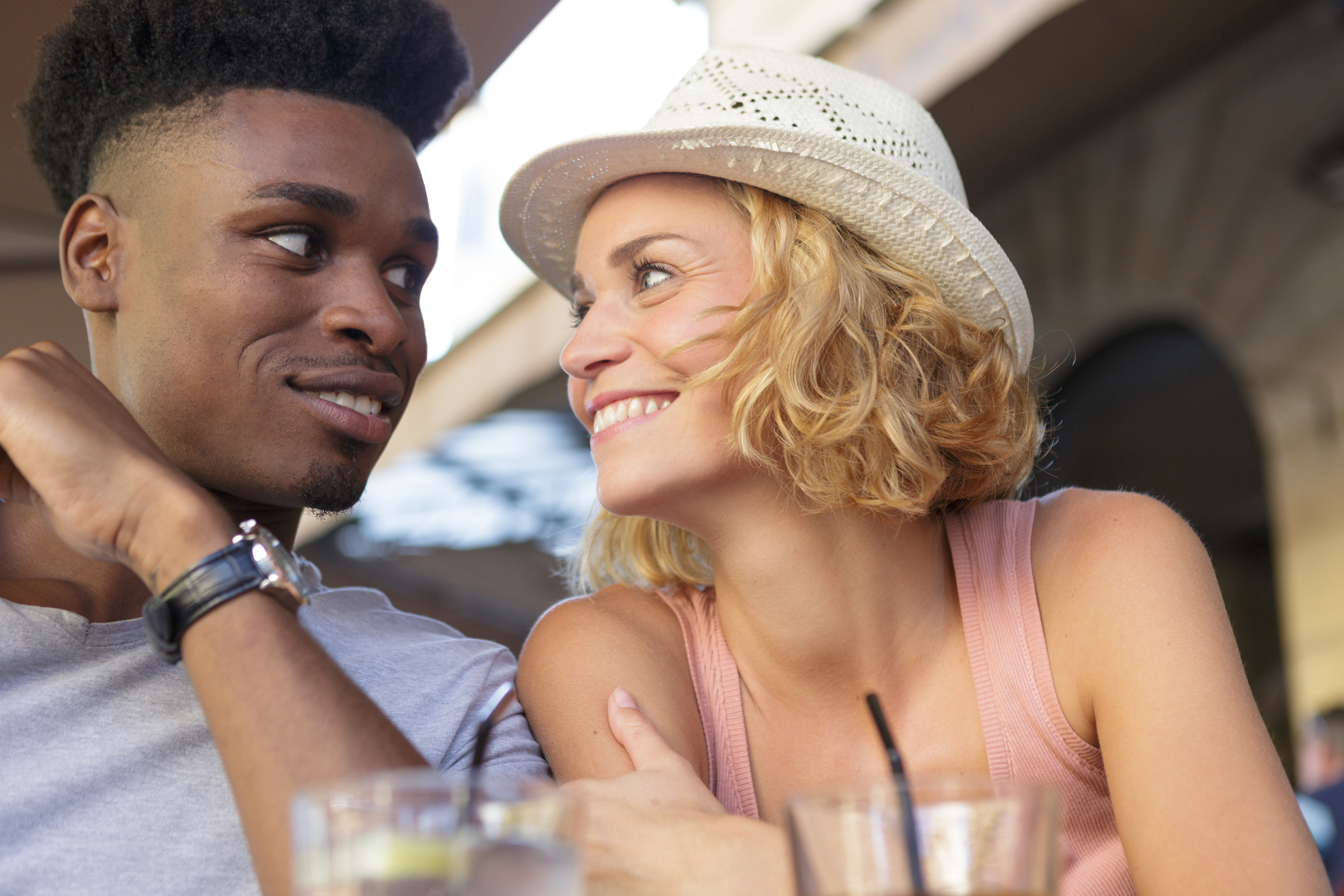 A young man and woman are smiling at each other while sitting closely at an outdoor table. The man has short curly hair and is wearing a gray shirt. The woman has a short blonde hairstyle, wears a white hat, and a pink tank top. Drinks are visible in front of them.