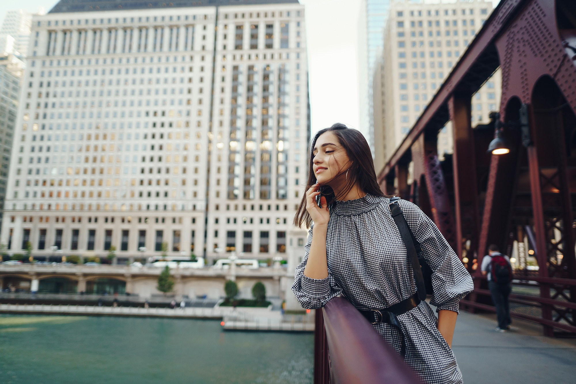 A woman in a black and white checkered dress leans against a railing on a bridge, gazing out over a river with large buildings in the background. She appears relaxed and content, enjoying the urban scenery on a sunny day.