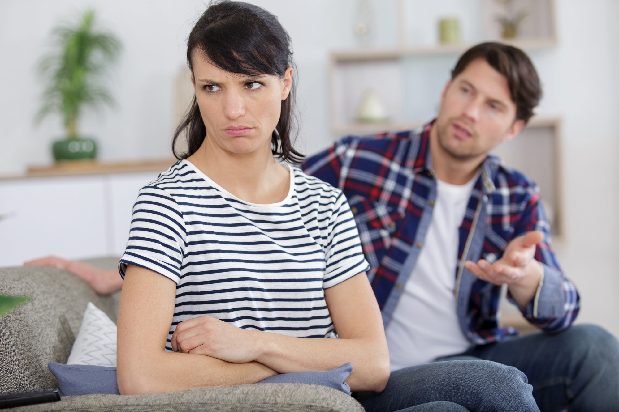 A woman with dark hair, wearing a striped shirt, sits on a couch with her arms crossed and an unhappy expression. A man with dark hair, wearing a plaid shirt, sits behind her, gesturing as if explaining or arguing. The room has a light, homey atmosphere.