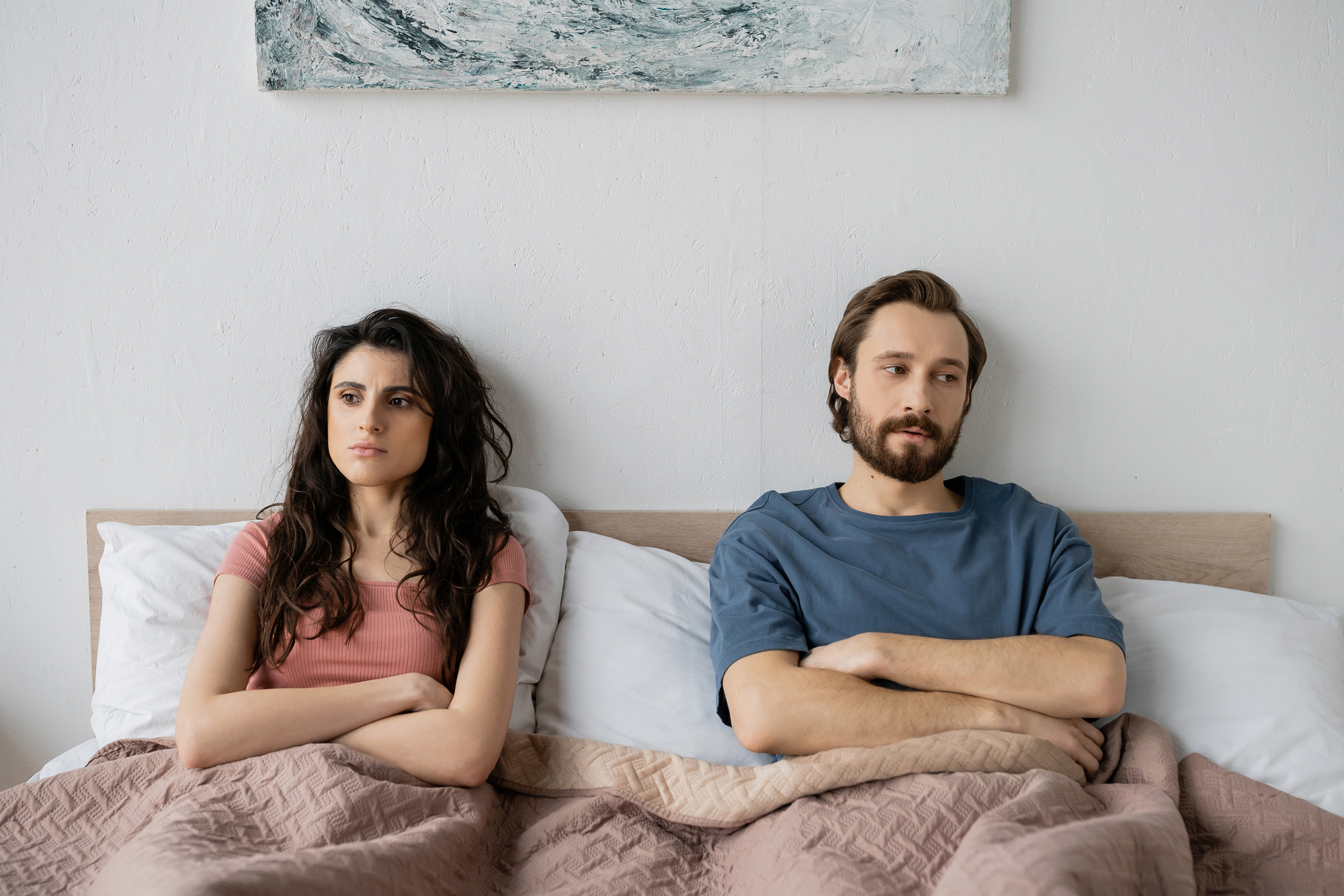 A man and woman are sitting in bed under a beige blanket, looking away from each other with serious expressions. The woman is wearing a pink shirt, and the man is wearing a blue shirt. A painting with a wave motif hangs on the wall behind them.