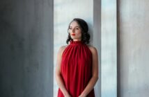 A woman with shoulder-length dark hair stands by a tall window, gazing thoughtfully into the distance. She is wearing a sleeveless, red halter-neck dress. The background is minimalist with light-grey textured walls.