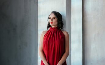 A woman with shoulder-length dark hair stands by a tall window, gazing thoughtfully into the distance. She is wearing a sleeveless, red halter-neck dress. The background is minimalist with light-grey textured walls.