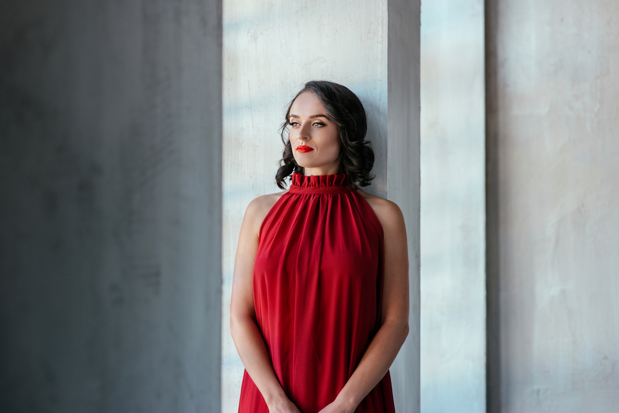 A woman with shoulder-length dark hair stands by a tall window, gazing thoughtfully into the distance. She is wearing a sleeveless, red halter-neck dress. The background is minimalist with light-grey textured walls.