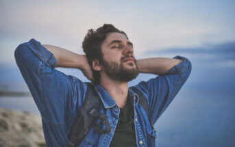 A man with a beard wearing a denim shirt and a backpack stands outdoors near the sea, stretching with his hands behind his head and eyes closed, appearing relaxed and content. The background features a cloudy sky and distant water.