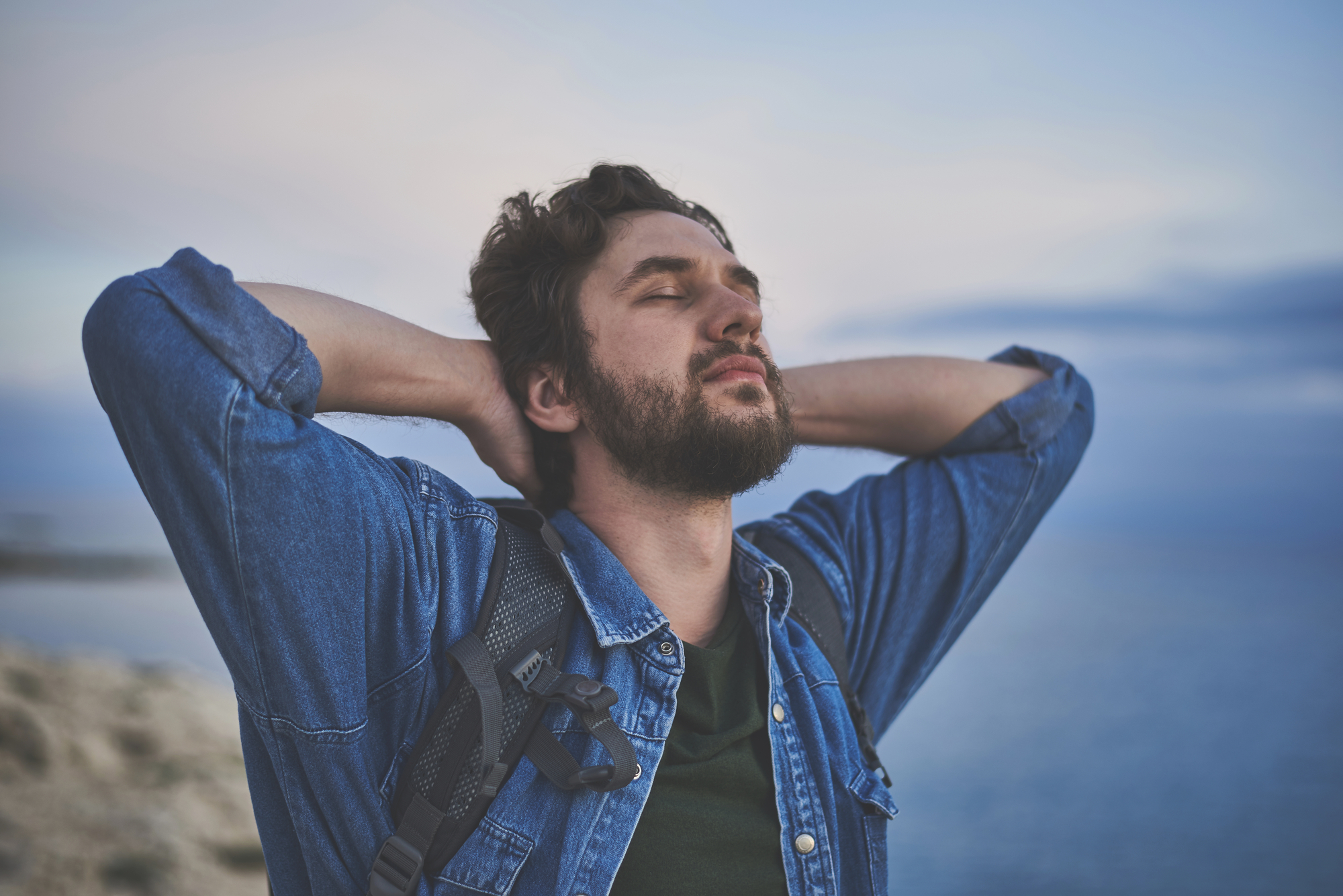 A man with a beard wearing a denim shirt and a backpack stands outdoors near the sea, stretching with his hands behind his head and eyes closed, appearing relaxed and content. The background features a cloudy sky and distant water.