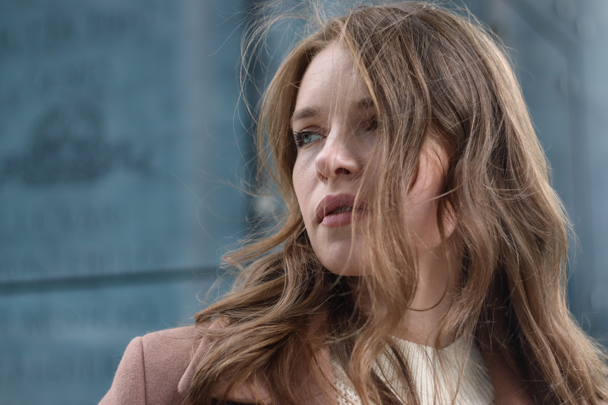 A woman with long, wavy brown hair is gazing thoughtfully to the side. She is wearing a light-colored sweater and a brown coat. The background appears to be an outdoor urban setting with blurred text visible on a wall.