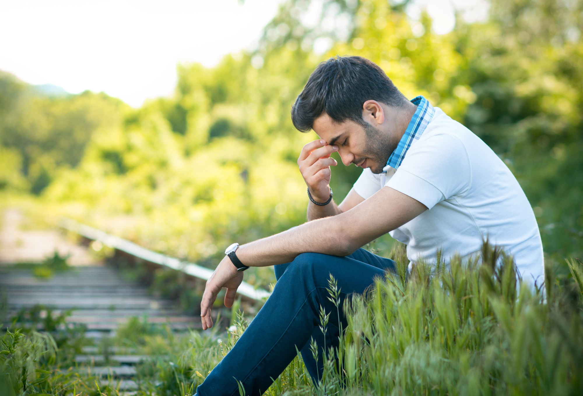 A man wearing a white shirt and blue pants sits on the ground next to overgrown railway tracks, holding his head and appearing distressed. The background features lush green foliage and blurred trees.