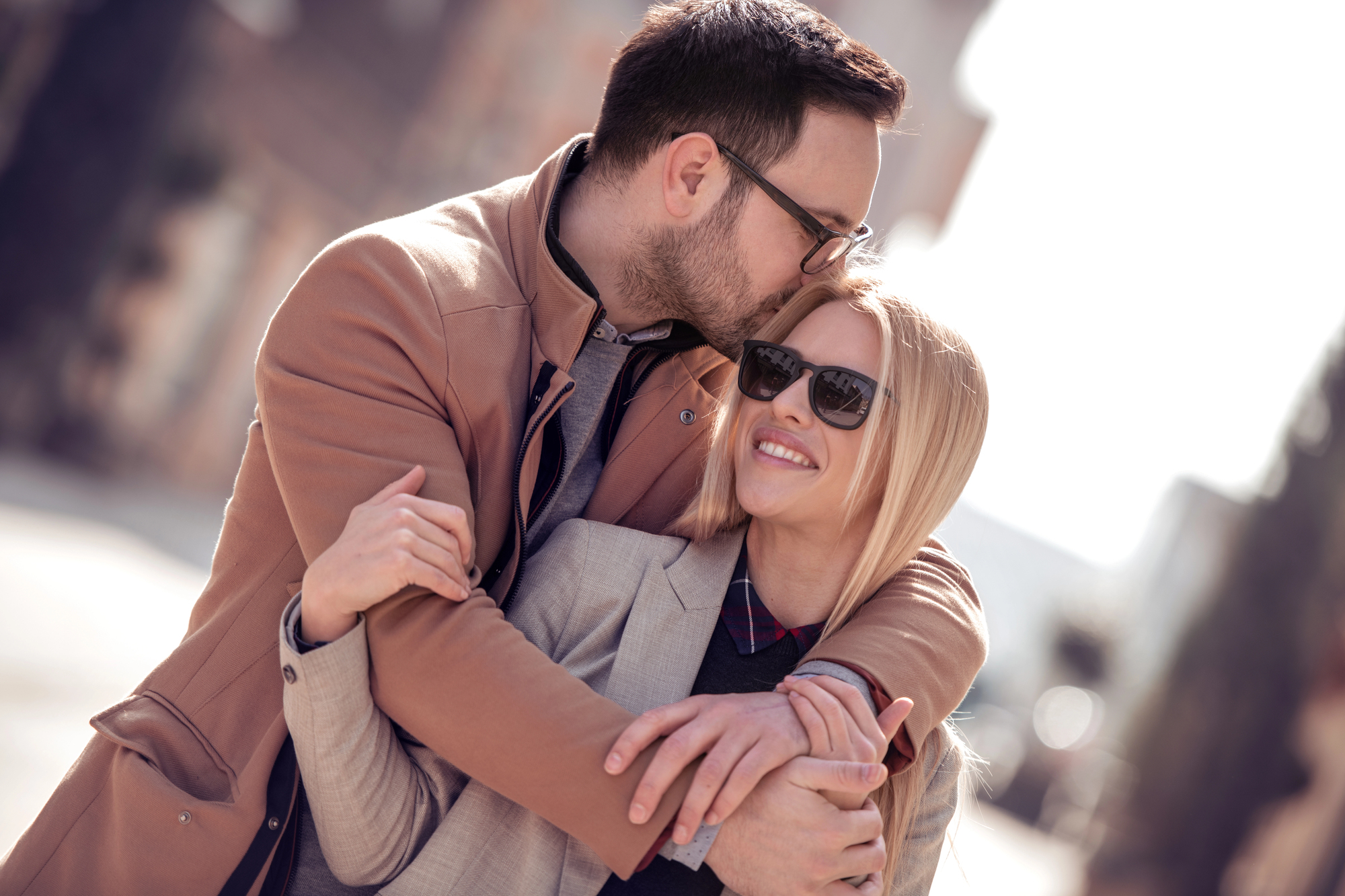 A man with dark hair and glasses kisses the forehead of a blonde woman wearing sunglasses. They are both smiling and dressed in casual coats. The woman has a light-colored coat while the man is in a brown coat. They appear to be outdoors on a sunny day.