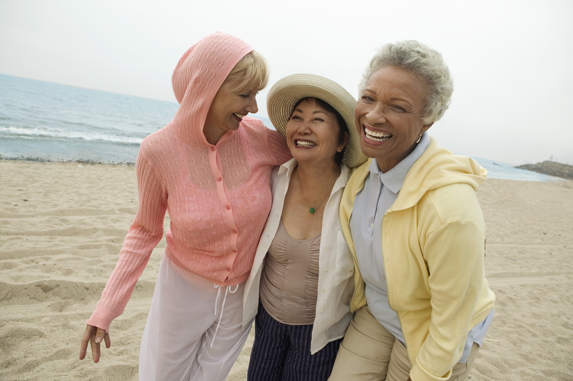 Three elderly women are smiling and walking arm-in-arm on a beach. One woman wears a pink hooded top, another wears a wide-brimmed hat and a beige top, and the third woman wears a yellow hooded top. The ocean is visible in the background.