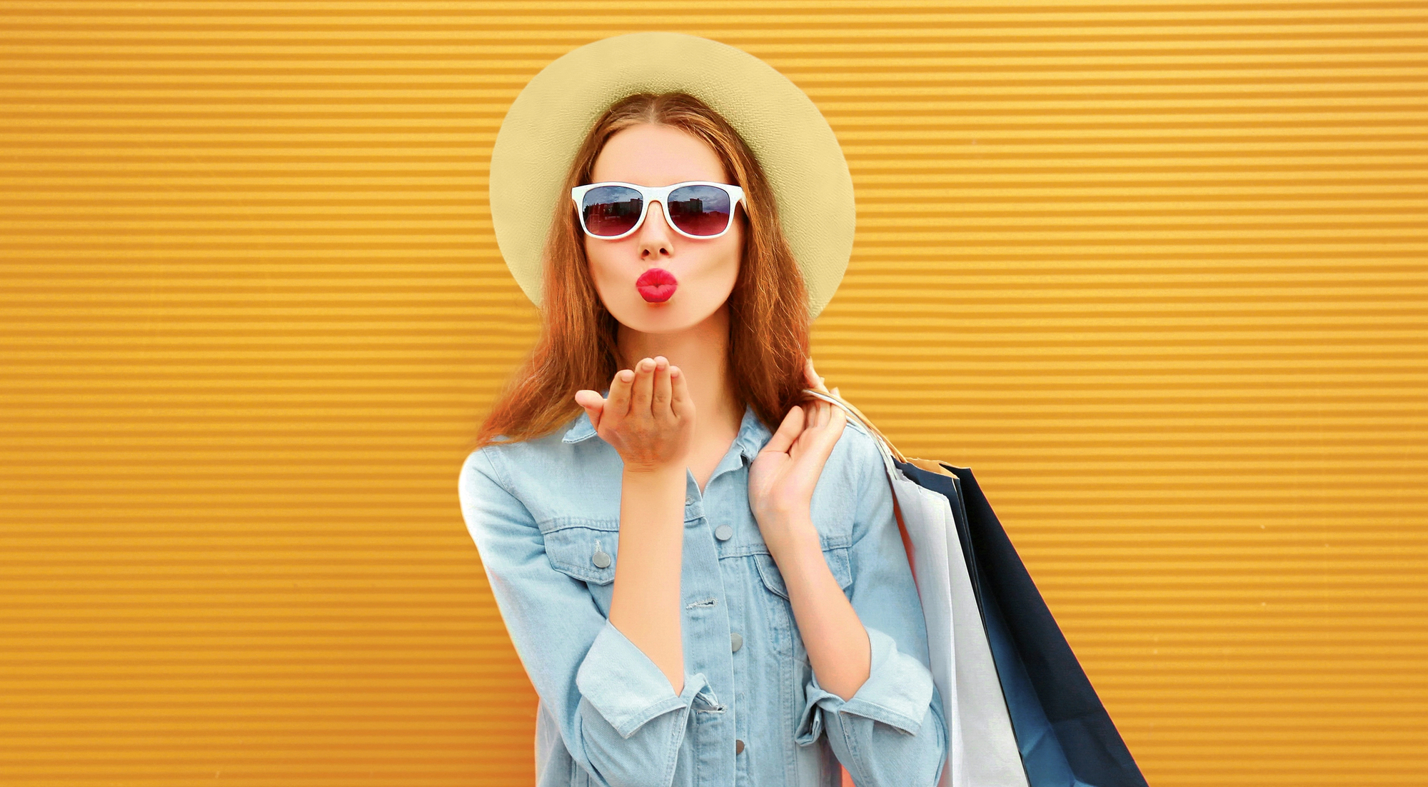 A woman with long brown hair wearing a wide-brimmed hat, white sunglasses, and a light denim jacket blows a kiss towards the camera. She holds several shopping bags and stands in front of a mustard yellow textured wall.