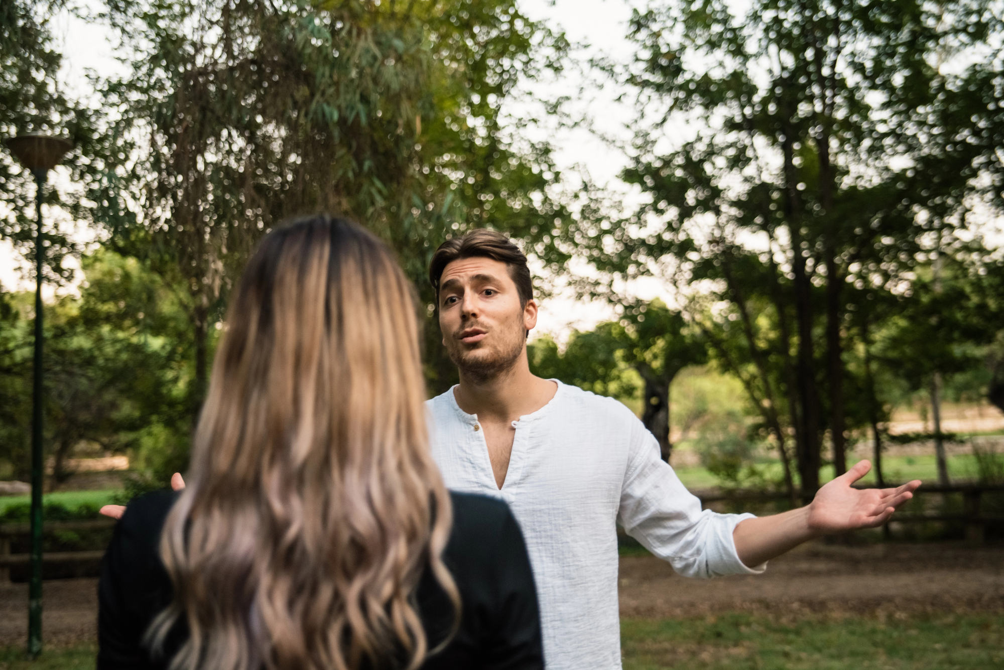 A man with short brown hair and a white shirt stands outdoors with his arms open, appearing to speak to a woman with long, blonde hair facing away from the camera. They are in a park-like setting, with trees and greenery in the background.