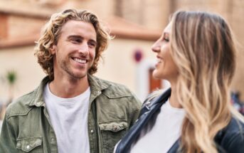 A man with wavy blonde hair wearing a green jacket and a white shirt smiles at a woman with long blonde hair wearing a black jacket and a white shirt. They are outside, with blurred buildings in the background.