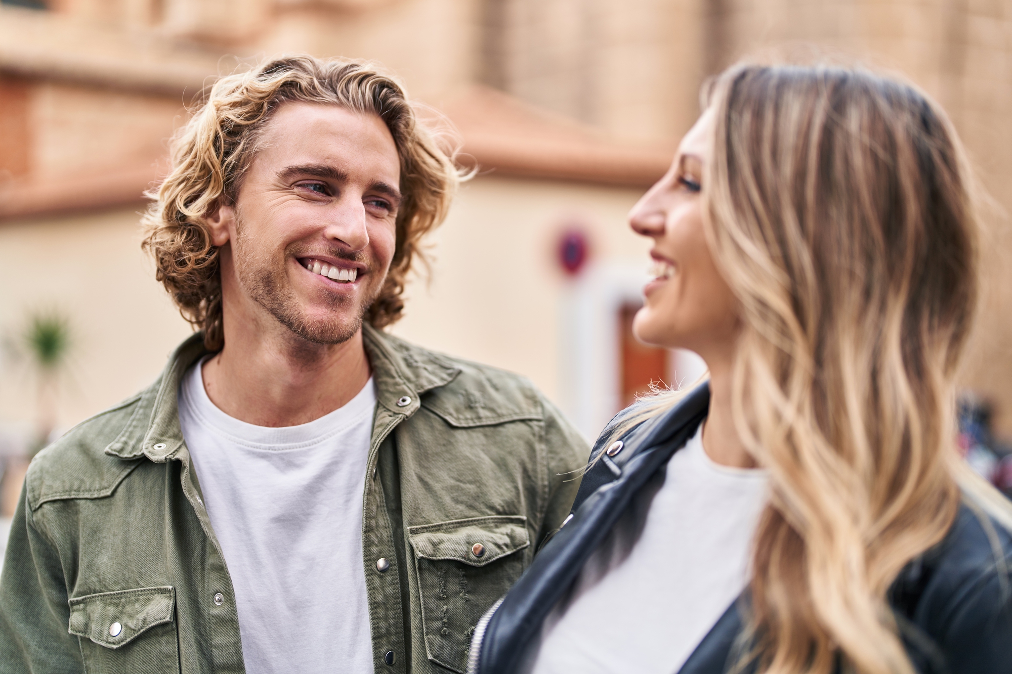 A man with wavy blonde hair wearing a green jacket and a white shirt smiles at a woman with long blonde hair wearing a black jacket and a white shirt. They are outside, with blurred buildings in the background.