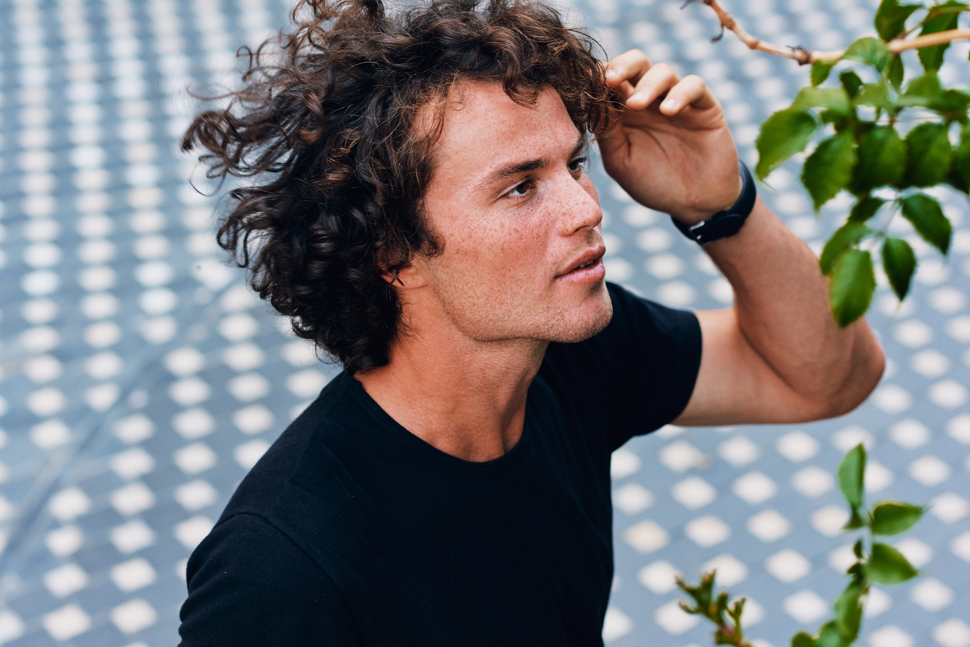 A person with curly hair looks into the distance while standing outdoors. They are wearing a black t-shirt and touching their head with one hand. In the foreground, there are some green leaves, and the background features a patterned surface.