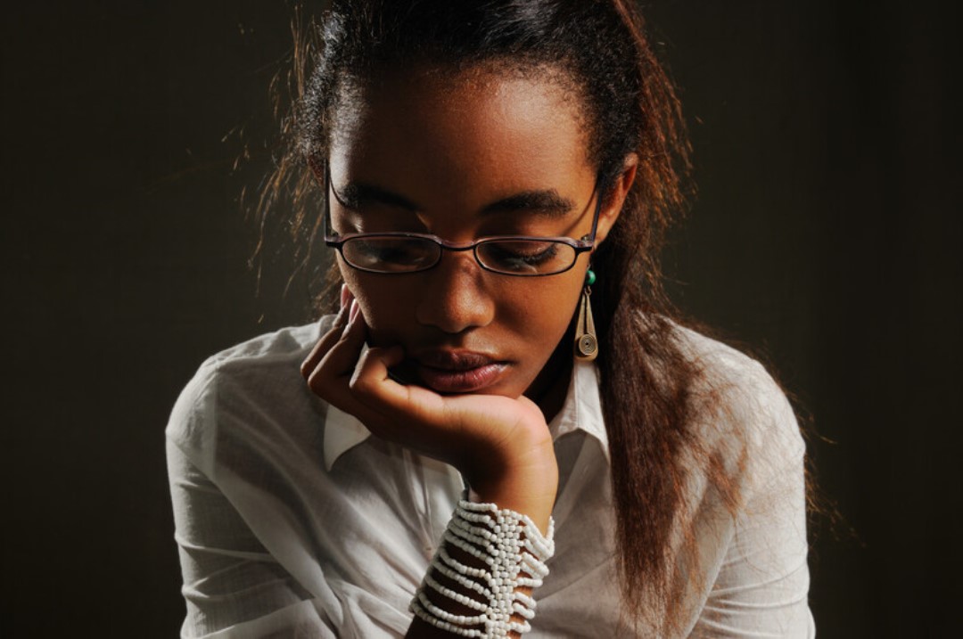 A young woman with glasses and long hair leans her head on her hand, deep in thought. She is wearing a white blouse and a beaded bracelet, with a dark background behind her.