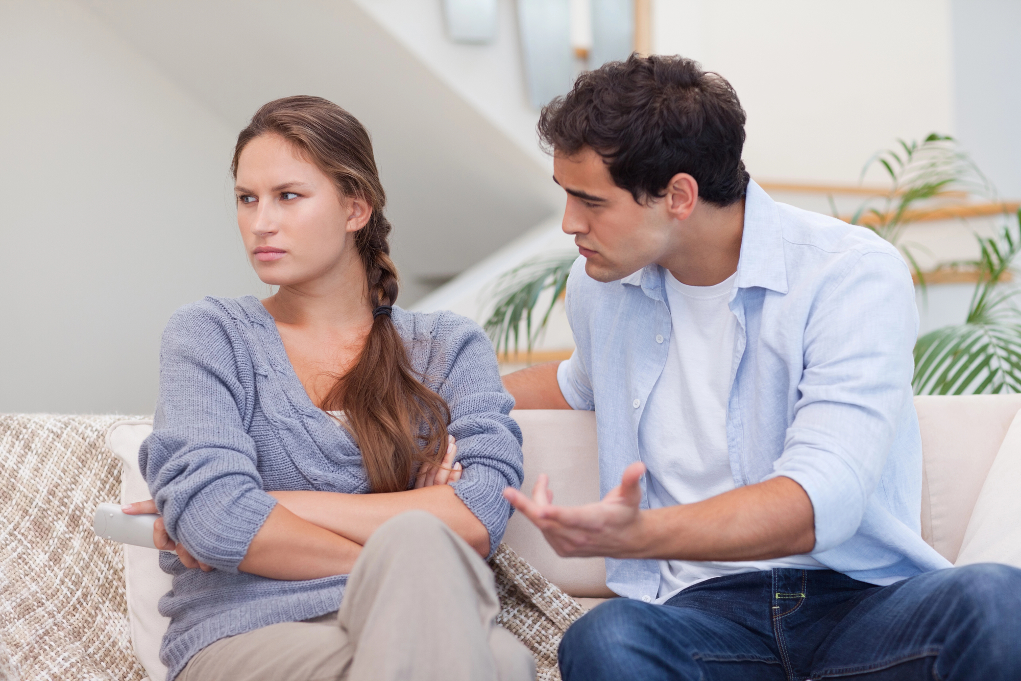 A young woman and man sit on a couch. The woman has her arms crossed and looks away, appearing upset or frustrated. The man is turned toward her, speaking with a concerned expression and gesturing with his hands, indicating he is trying to communicate.