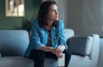 A person with shoulder-length brown hair, wearing a denim shirt over a striped top and black pants, sits on a gray couch holding a smartphone. They gaze thoughtfully into the distance with a serious expression. The background is blurred with a hint of green wall.