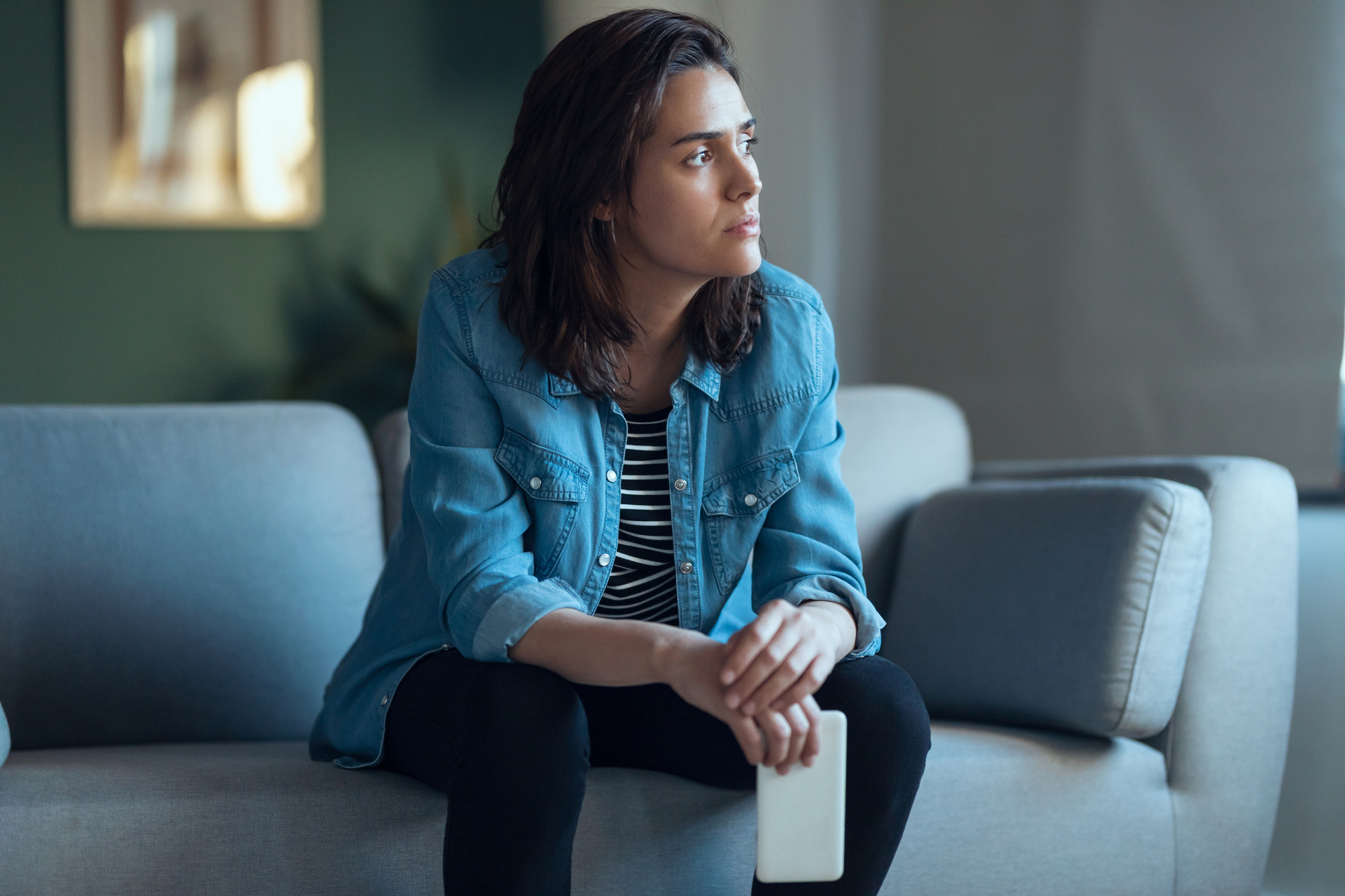 A person with shoulder-length brown hair, wearing a denim shirt over a striped top and black pants, sits on a gray couch holding a smartphone. They gaze thoughtfully into the distance with a serious expression. The background is blurred with a hint of green wall.