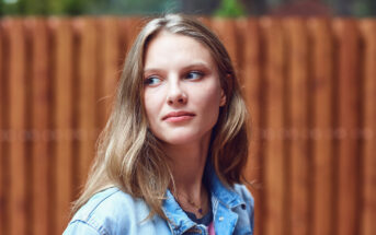 A young woman with long, light brown hair looks thoughtfully to her right. She is wearing a light blue denim jacket, and a wooden fence is visible in the blurry background. The image captures her in natural light, giving a serene and contemplative feel.