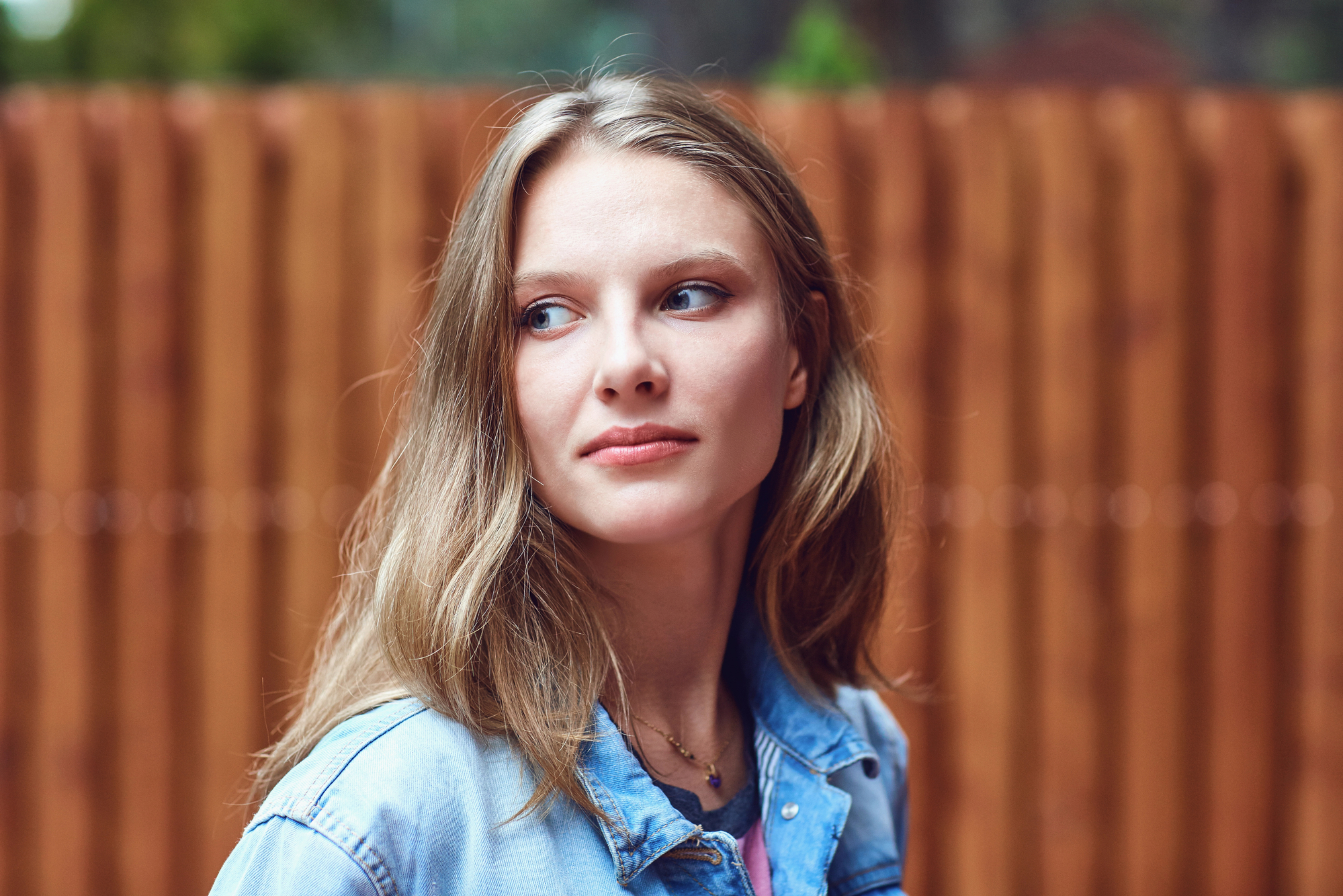 A young woman with long, light brown hair looks thoughtfully to her right. She is wearing a light blue denim jacket, and a wooden fence is visible in the blurry background. The image captures her in natural light, giving a serene and contemplative feel.