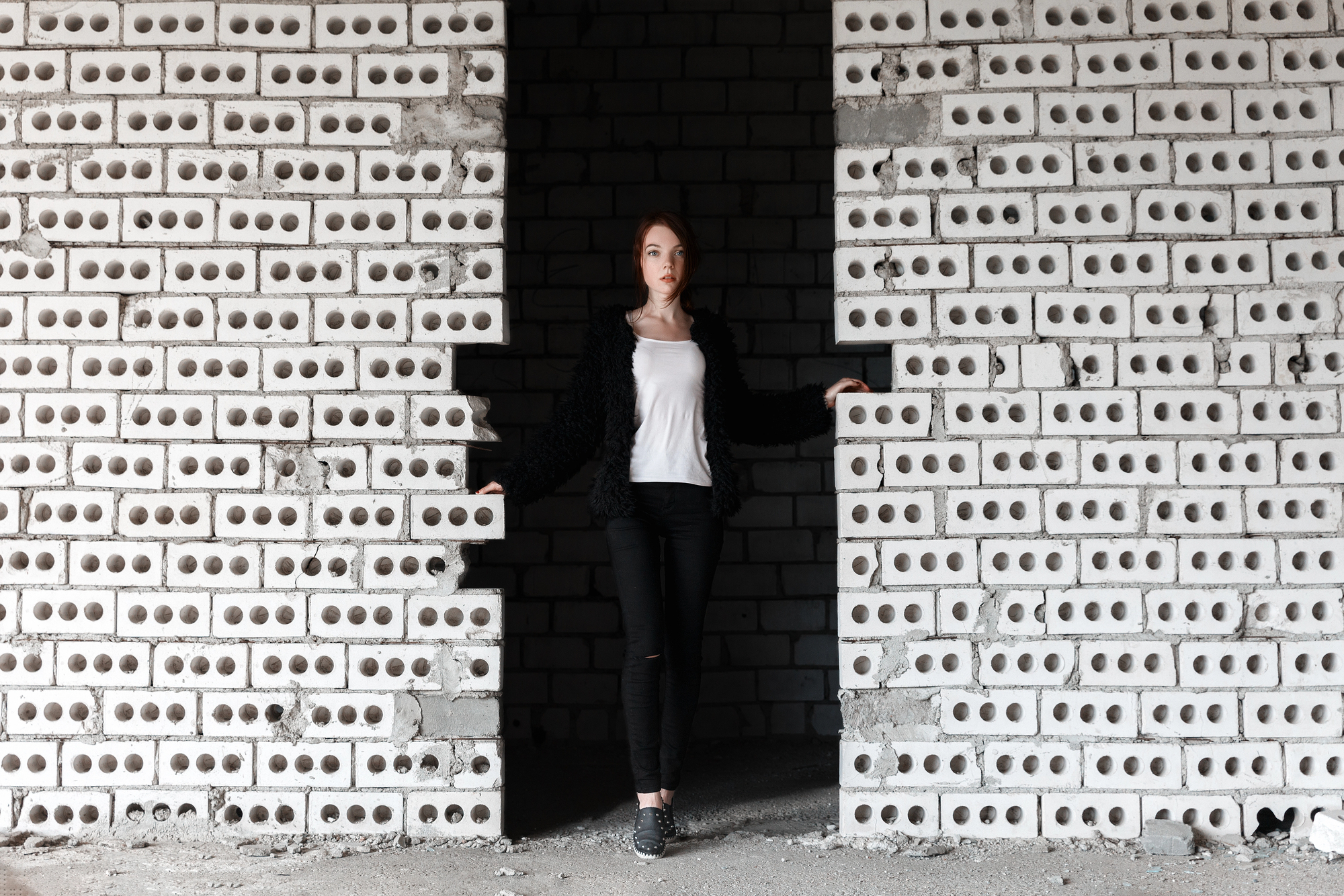 A person stands in a doorway of an unfinished brick wall. They are wearing a white shirt, black jacket, and black pants. Their hands are touching the sides of the doorway, and they are looking directly at the camera. The surrounding walls are made of hollow bricks.