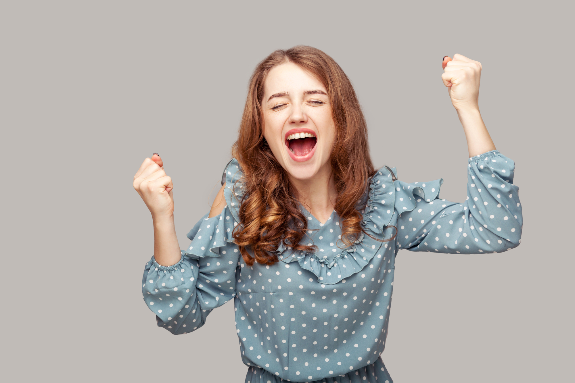 A young woman with long brown hair, wearing a blue polka dot blouse, is joyfully raising both fists in the air. Her eyes are closed, and she has a wide, happy smile on her face, conveying a sense of excitement and triumph. The background is plain gray.