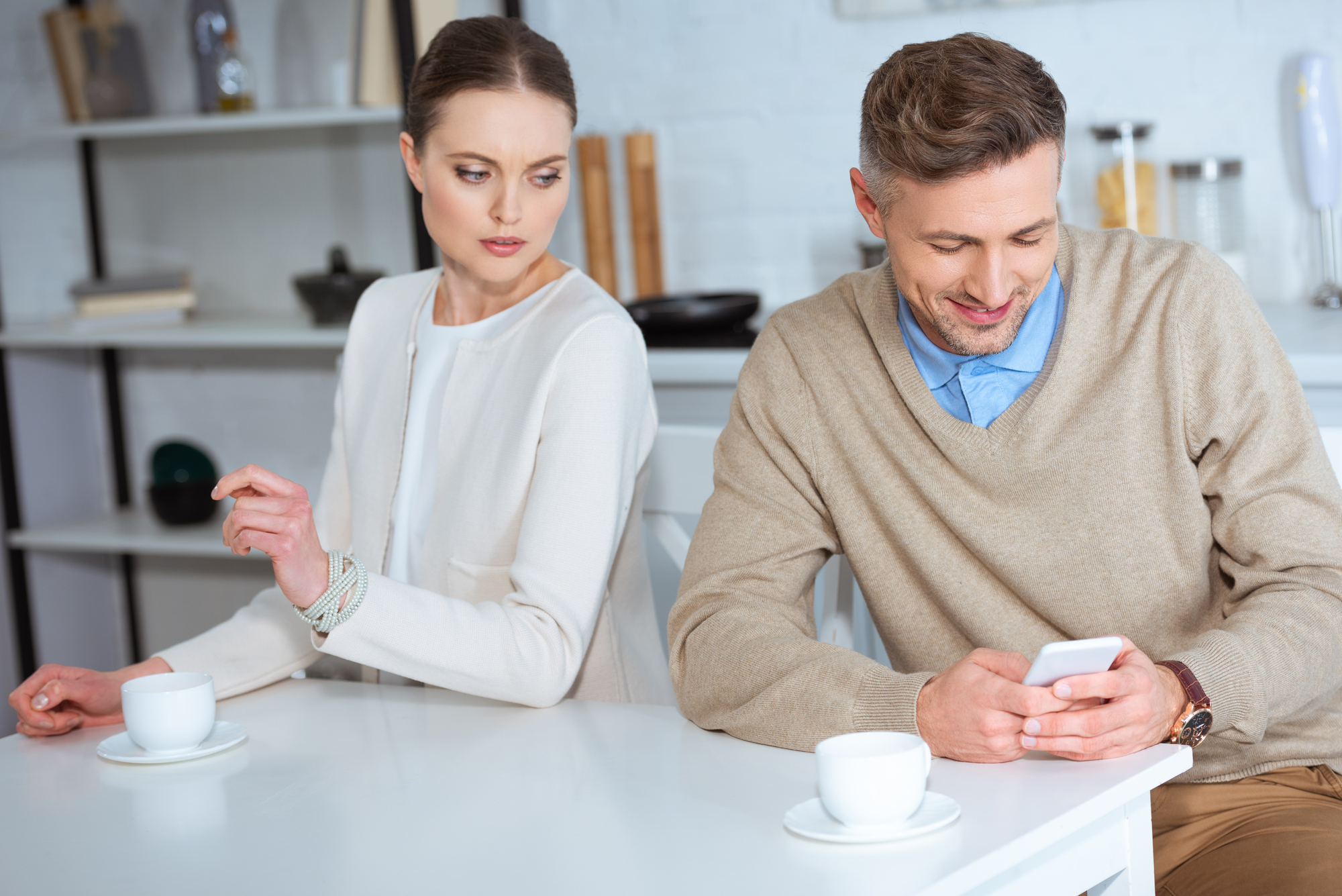 A woman in a white top looks over at a man in a beige sweater who is smiling while using his smartphone. They are sitting at a white table with coffee cups in an indoor setting. The woman appears curious or possibly suspicious.