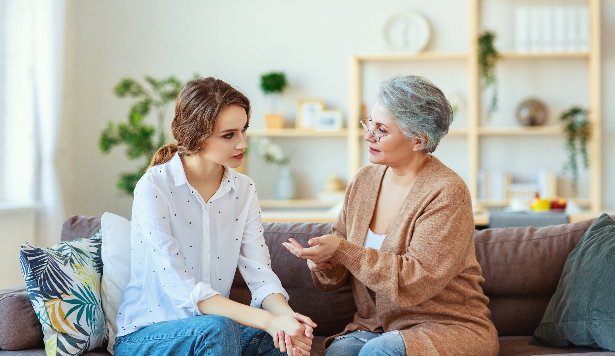 A young woman and an older woman are sitting on a couch having a serious conversation. The young woman has brown hair and is wearing a white polka dot shirt and jeans. The older woman has gray hair and is wearing a beige cardigan and glasses. Shelves with plants and decor are in the background.