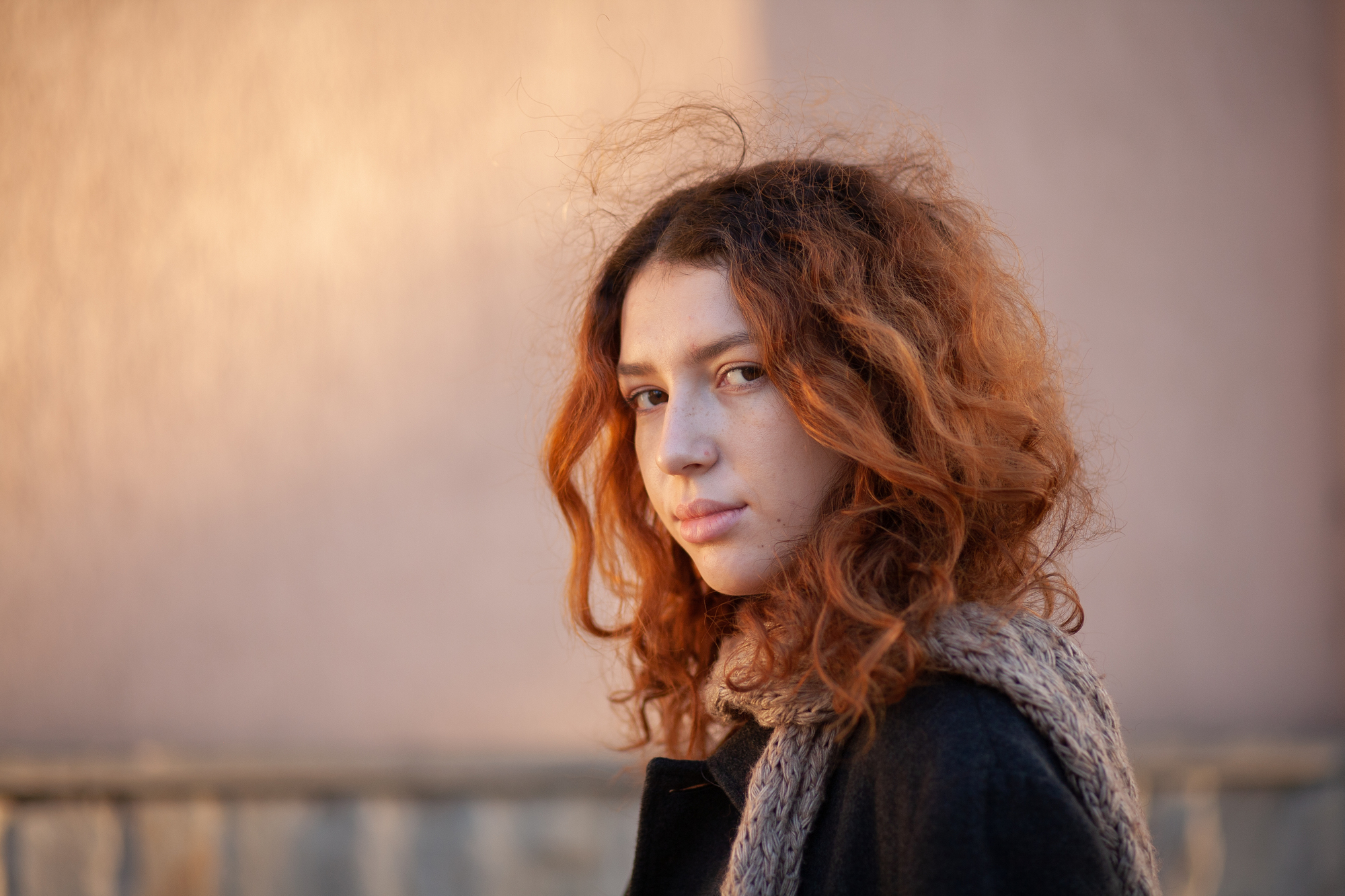 A woman with red curly hair stands outdoors, gazing intently at the camera. She wears a dark coat and a knitted scarf, with a soft, blurred background that suggests a late afternoon or early morning light. The atmosphere appears calm and contemplative.