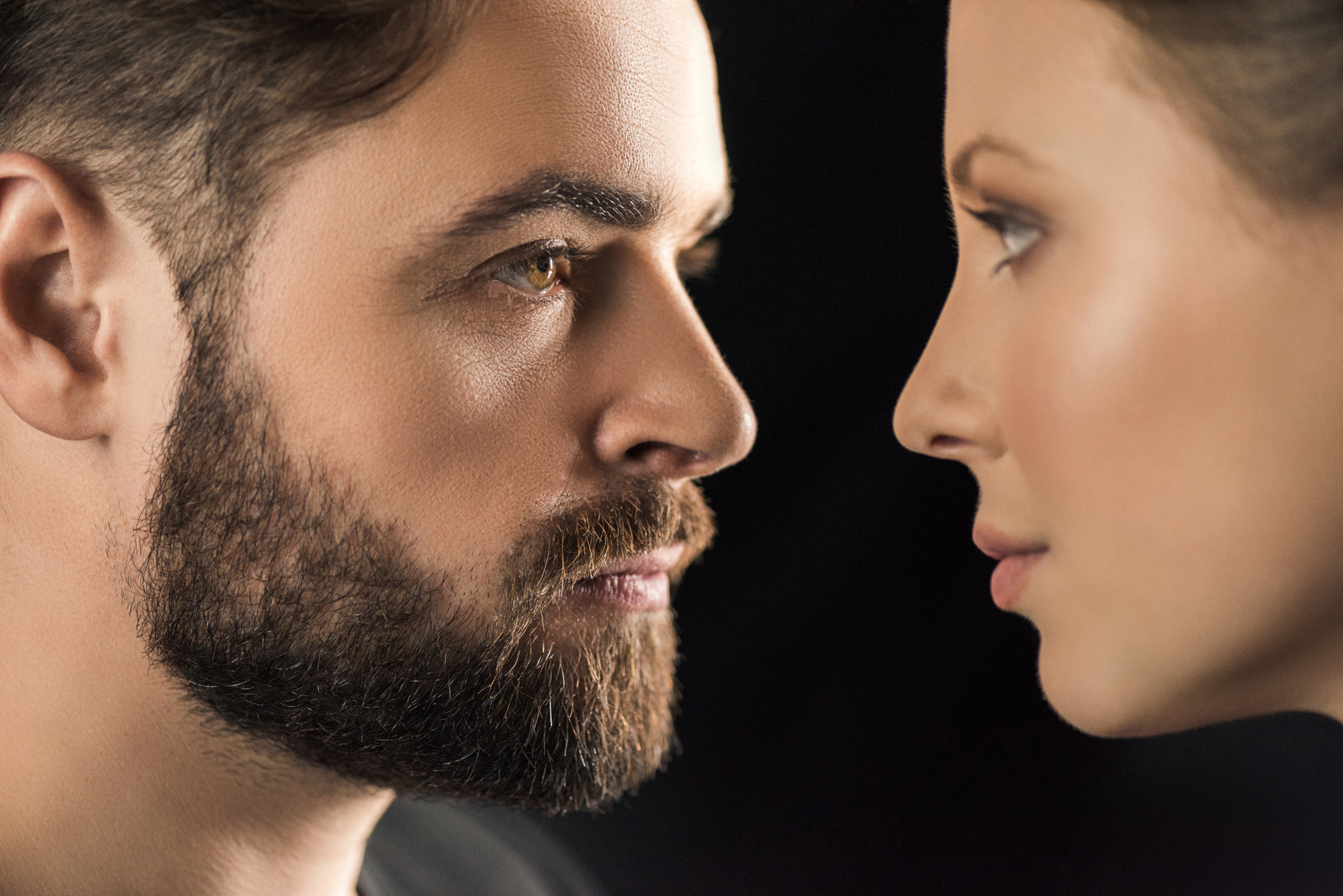 Close-up profile view of a man with a beard and a woman staring at each other against a black background. The man's face is on the left, and the woman's face is on the right, both looking intently at each other.