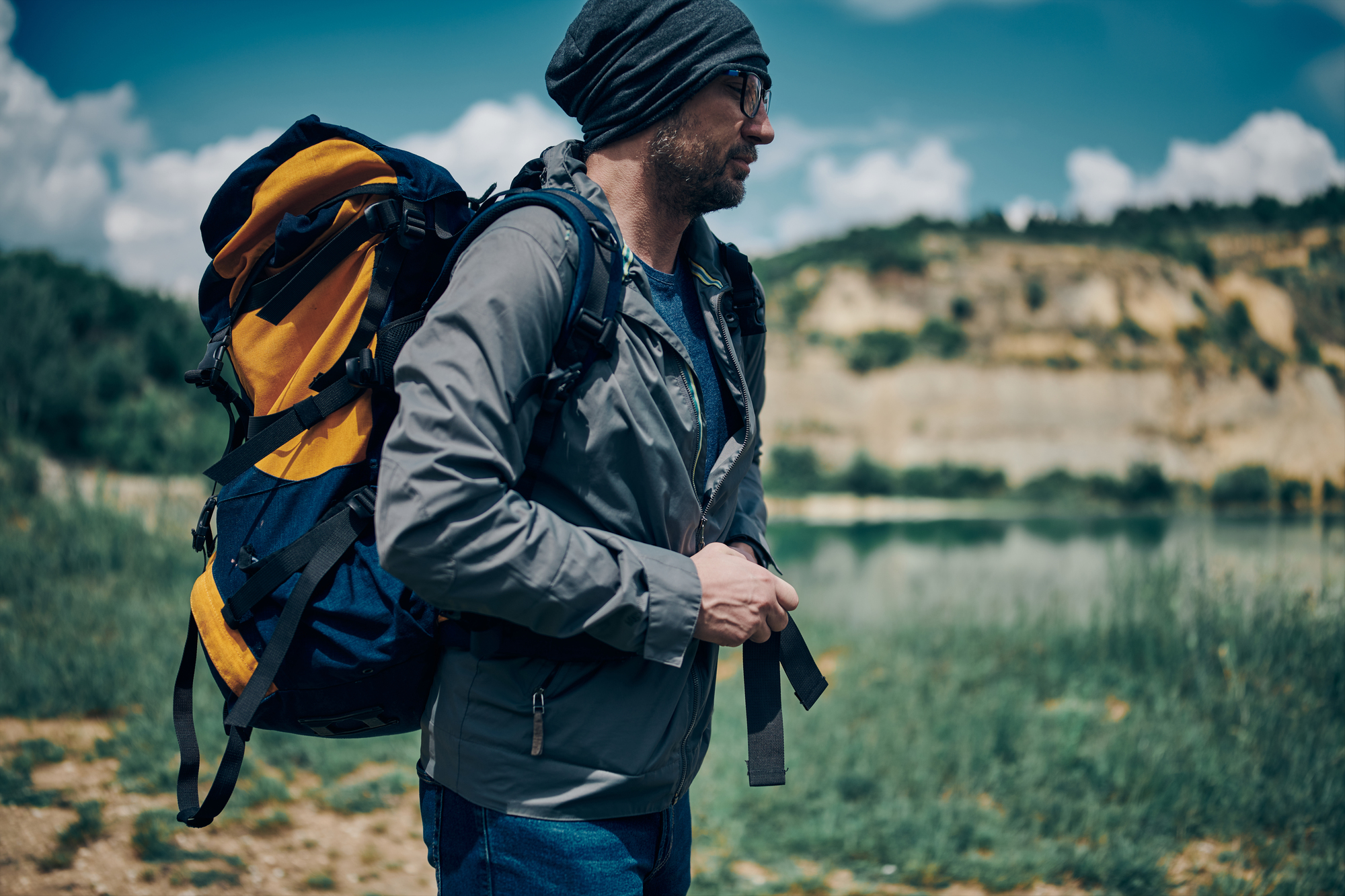 A person in a gray jacket and beanie walks outdoors while carrying a large backpack. They are in a natural setting with a lake and hills in the background, under a partly cloudy sky.