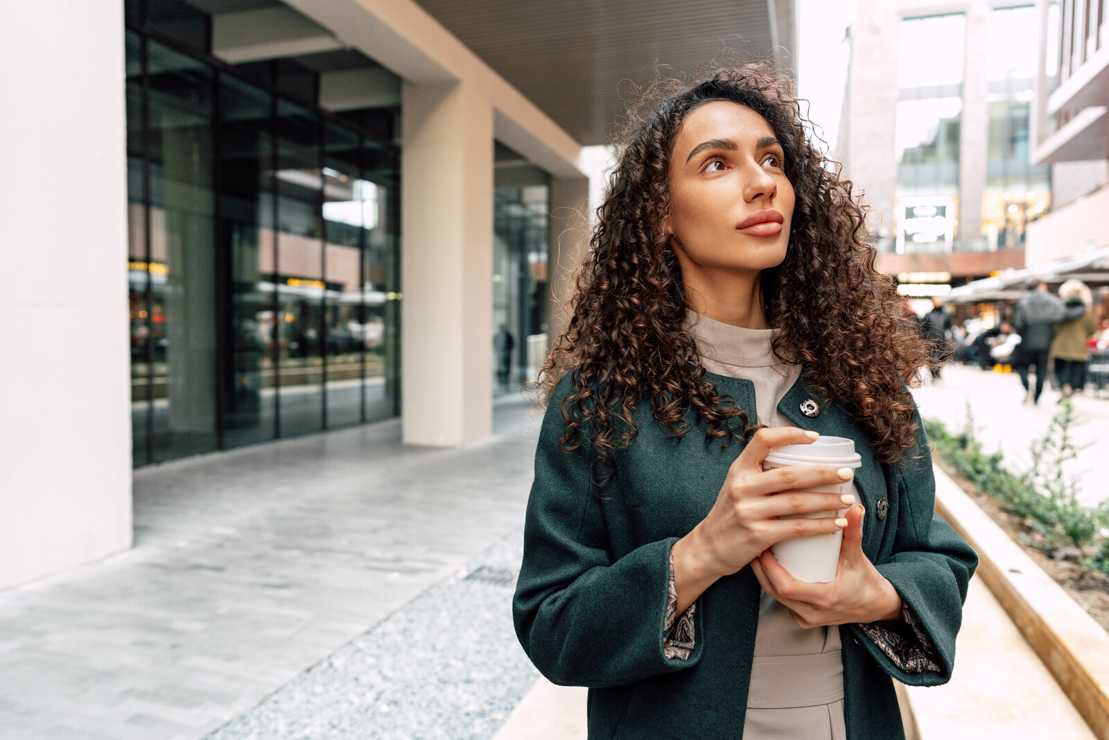 A woman with curly hair wearing a dark green coat is standing outside in an urban setting holding a white disposable coffee cup. She is looking thoughtfully into the distance. There are modern buildings and people walking in the background.