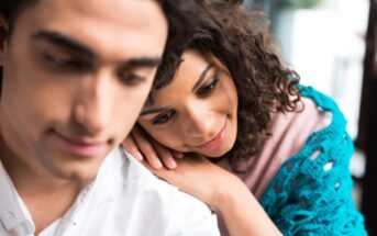 A close-up of a couple. The woman with curly hair is leaning her head on the man's shoulder, smiling gently. The man, also with dark hair, is looking downward with a soft expression. They are indoors, and the background is softly blurred.