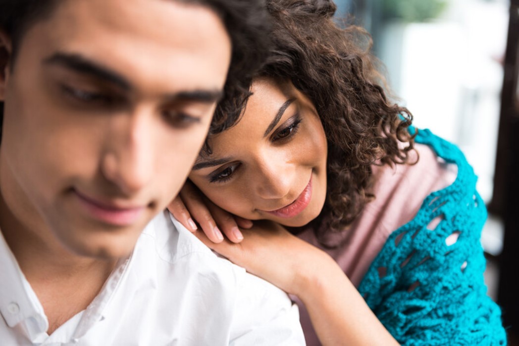 A close-up of a couple. The woman with curly hair is leaning her head on the man's shoulder, smiling gently. The man, also with dark hair, is looking downward with a soft expression. They are indoors, and the background is softly blurred.