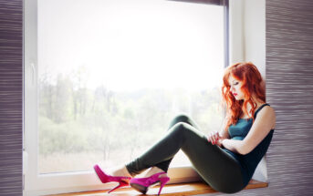 A woman with long red hair is sitting on a windowsill. She is wearing a black tank top, green pants, and bright pink high heels. She gazes out the window with a thoughtful expression. The background shows a blurred, green, outdoor landscape through the window.