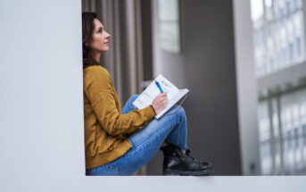 A woman with shoulder-length dark hair, wearing a mustard yellow jacket and blue jeans, sits in a windowsill holding an open book and a pen. She appears thoughtful, gazing upwards while surrounded by a modern, minimalist indoor setting.