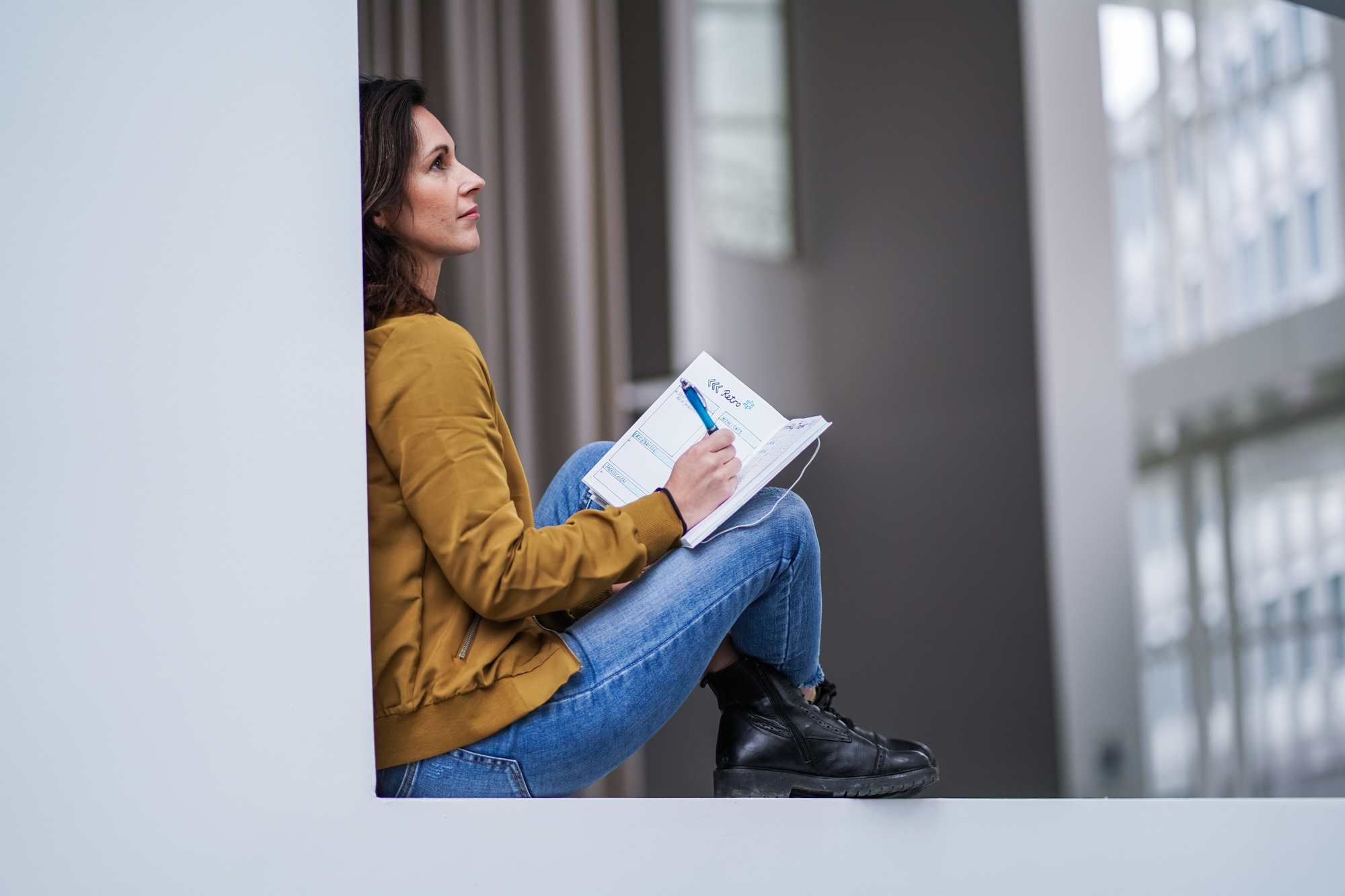 A woman with shoulder-length dark hair, wearing a mustard yellow jacket and blue jeans, sits in a windowsill holding an open book and a pen. She appears thoughtful, gazing upwards while surrounded by a modern, minimalist indoor setting.