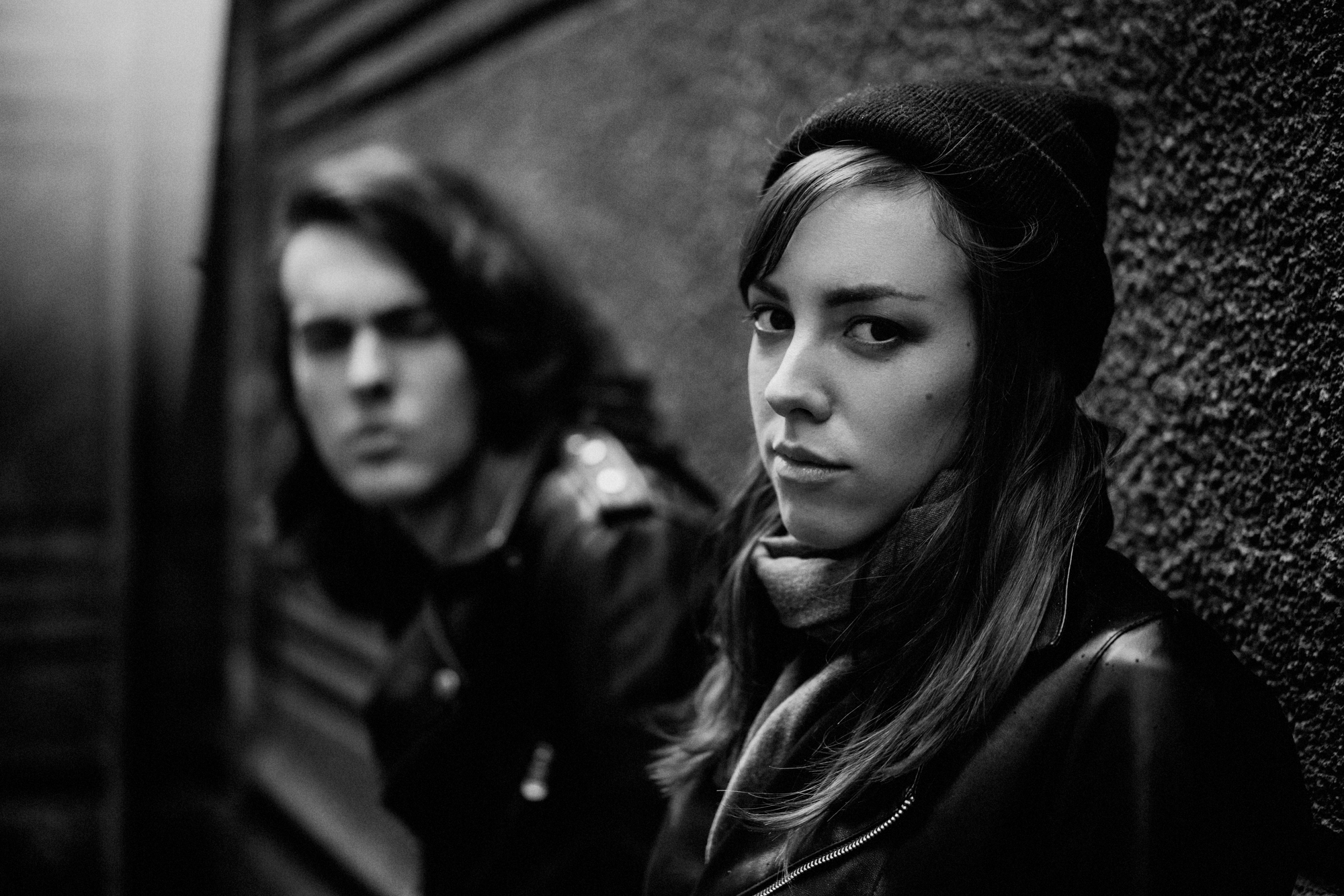 Black and white photo of two individuals leaning against a textured wall. The person in the foreground, wearing a beanie and scarf, looks directly at the camera with a neutral expression. The person in the background is slightly out of focus. Both are dressed in dark clothing.