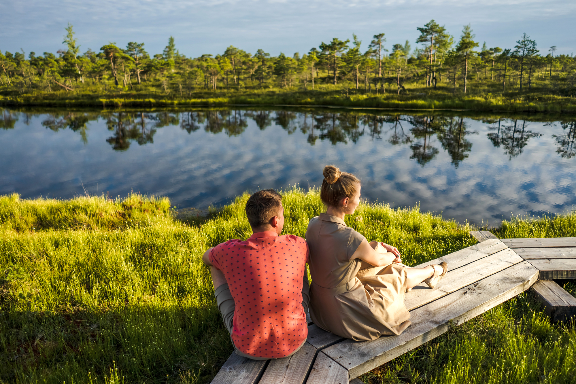 A man in a red polka-dot shirt and a woman in a beige dress sit on a wooden platform by a serene lake. They sit closely, gazing at the calm water reflecting the sky and surrounding lush greenery, under a blue sky with scattered clouds.