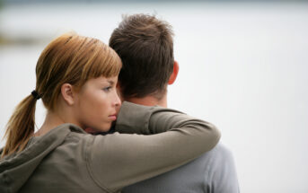 A woman with light brown hair rests her head on the shoulder of a man with short brown hair. They are standing close to each other, with the woman looking off into the distance. The background is blurry and appears to be an outdoor setting, possibly near water.